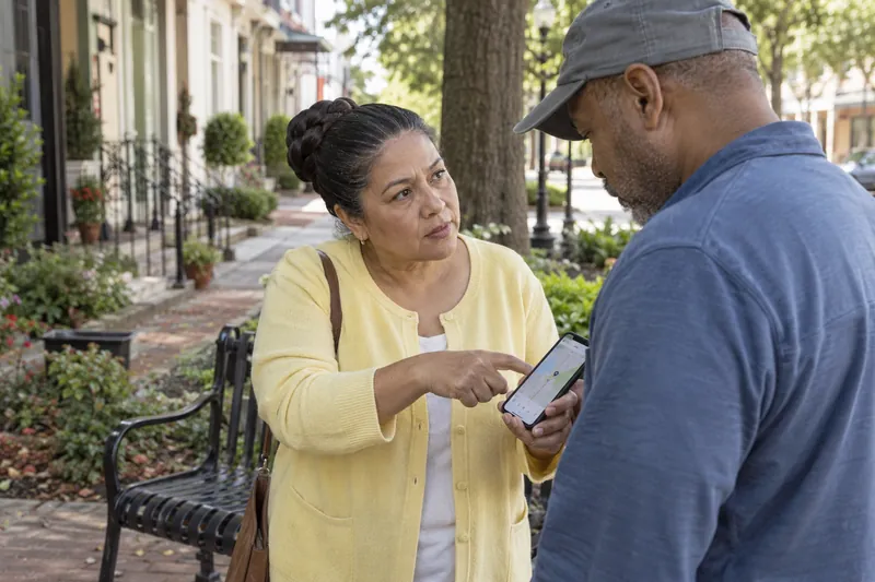 A visitor asking a companion for directions while pointing at a phone map near a public bench