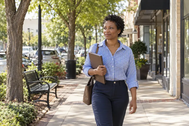 A visitor arriving on foot along a tree-lined neighborhood commercial street, carrying a slim folder