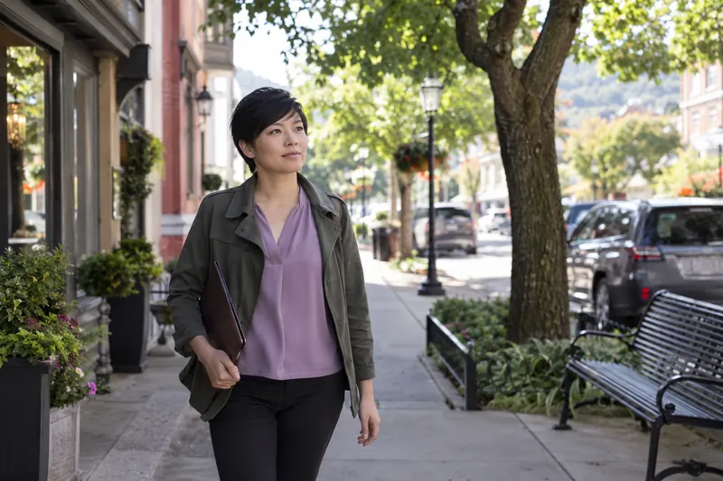 A visitor arriving on foot along a tree-lined neighborhood commercial street, carrying a slim folder