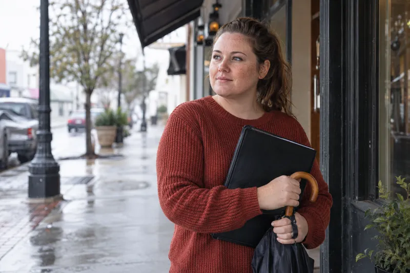 A visitor standing under a storefront awning with a closed umbrella and appointment folder